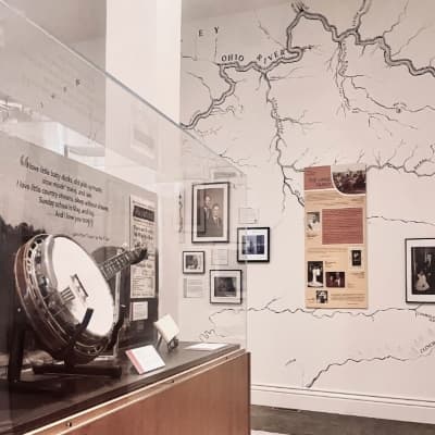 A gallery view inside the Carriage House: a banjo on display in a glass case beside a wall map of the Ohio River watershed and framed historic photographs.